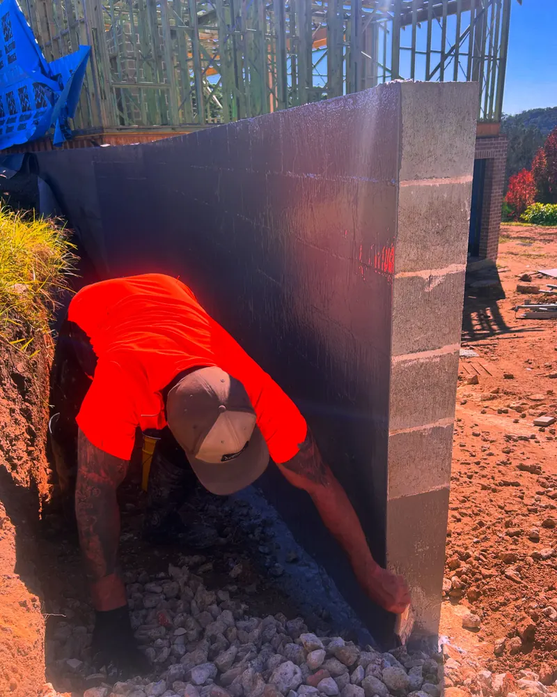 Marty brushing black membrane onto a block retaining wall above drainage gravel
